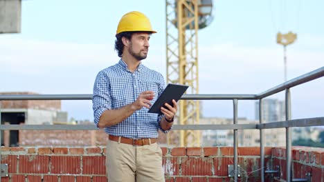 Caucasian-young-good-looking-man-architect-in-the-hardhat-standing-at-the-roof-of-the-building-site-using-tablet-device,-tapping-and-scrolling.